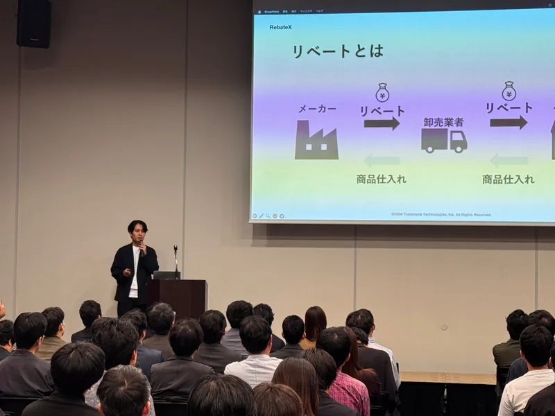 ベルサール六本木の会場で、「リベートとは」のスライドを背にマイクを持って話す玉城