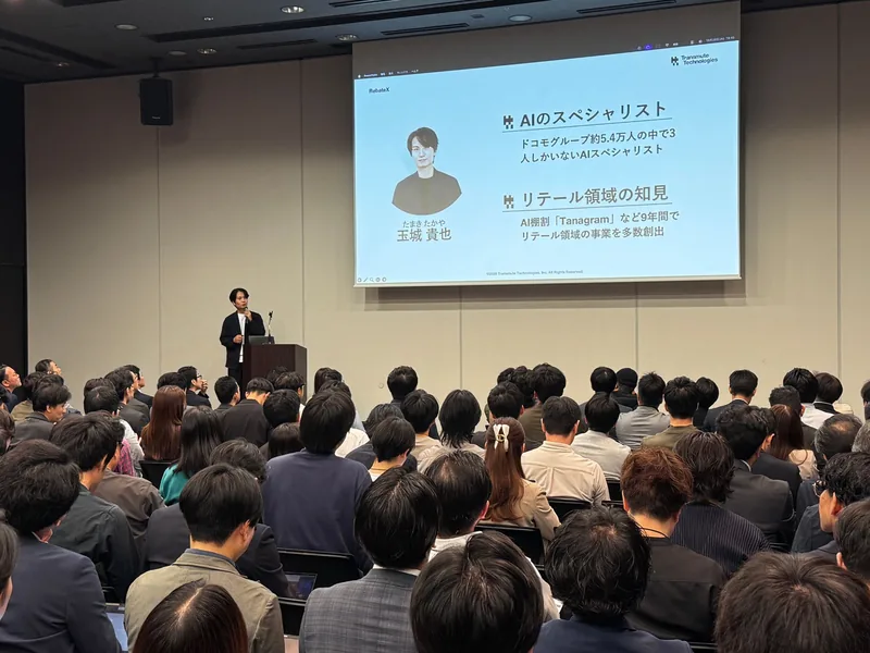 満席の会場で、登壇者紹介のスライドを背に話す玉城